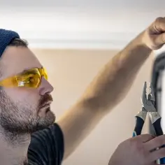 Elektryk w okularach ochronnych i czapce z wąsami, trzymający kombinerki, podłącza przewody w puszce. Ważne jest, z której strony faza w gniazdku.