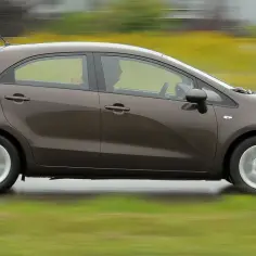 Brązowy Kia Rio w ruchu. Kierowca w środku, zielony krajobraz w tle. Wiele osób szuka kia rio opinie przed zakupem.
