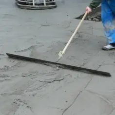 Pracownik w niebieskich ogrodniczkach wygładza świeży beton długą łopatą. Trwa wygrzewanie posadzki.