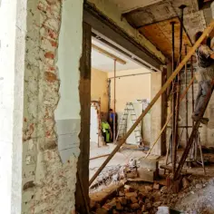Budowlaniec na drabinie pracuje przy podciągu stropowym. W tle widać gruz i inne materiały budowlane.