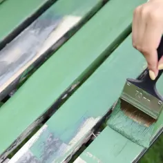 Ręka maluje zieloną farbą drewniane meble ogrodowe. To świetny sposób, by odnowić stare krzesło i dowiedzieć się, czym pomalować meble ogrodowe.