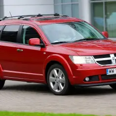 Czerwony Dodge Journey 3.6 z relingami dachowymi jedzie po brukowanej drodze obok nowoczesnego budynku.