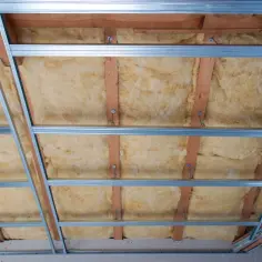 Ocieplenie stropu betonowego w trakcie prac. Widoczna wełna mineralna między drewnianymi belkami, pod profilem metalowym do sufitu podwieszanego.