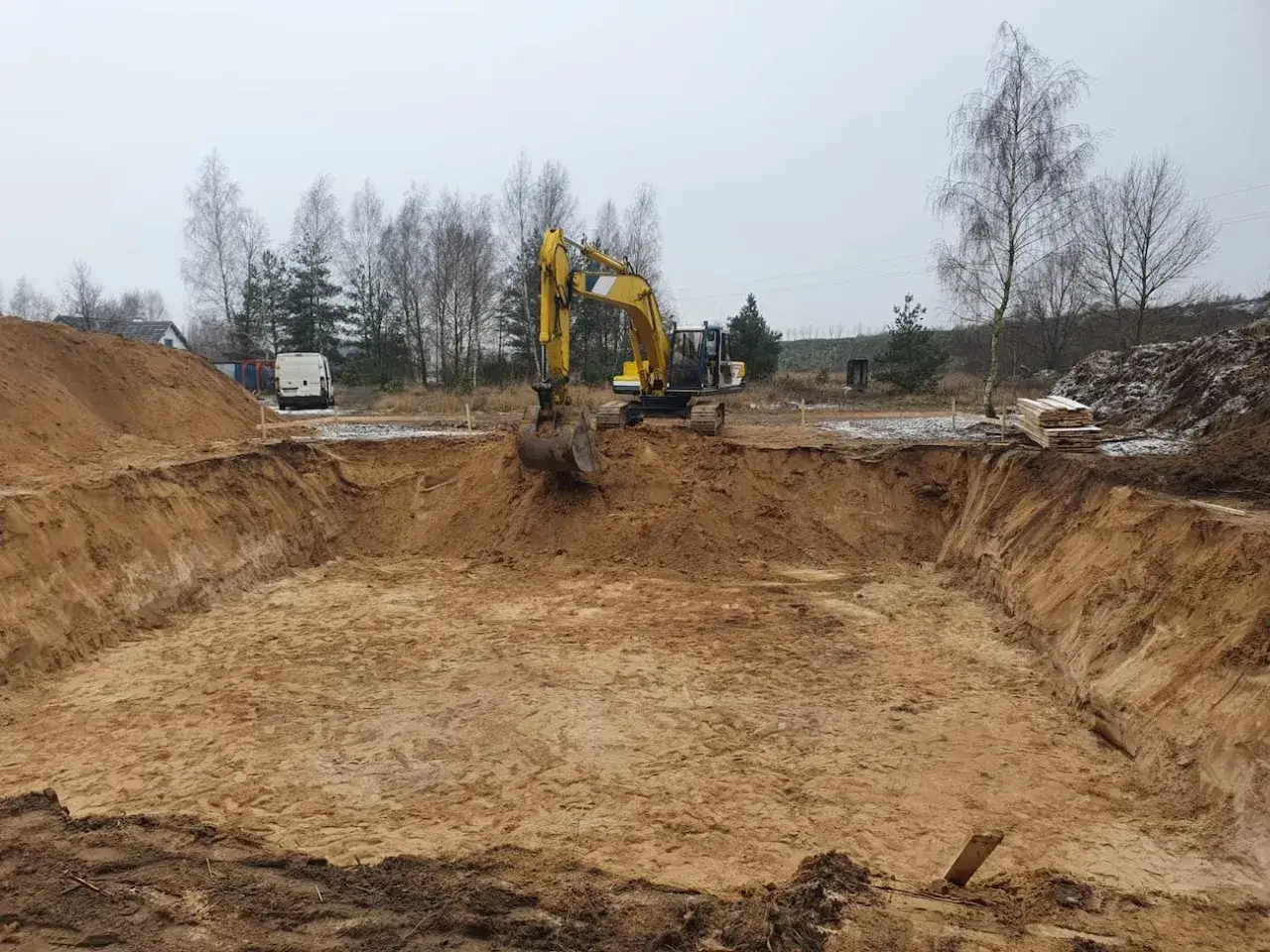 Koparka pracuje na budowie, tworząc wykop. Piasek i ziemia tworzą kurzawki wokół.
