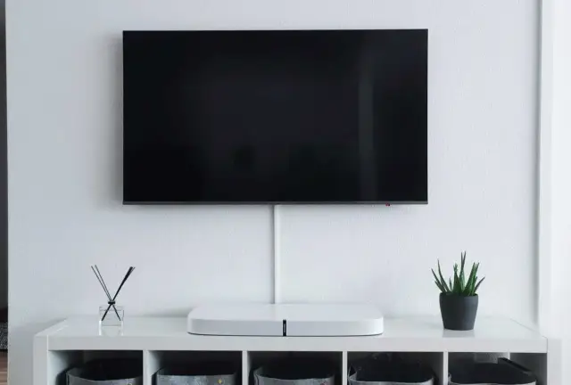 Minimalistyczny salon z telewizorem zamontowanym na ścianie. Na białej komodze stoi biały soundbar i mała roślinka. Idealna na jakiej wysokości telewizor.